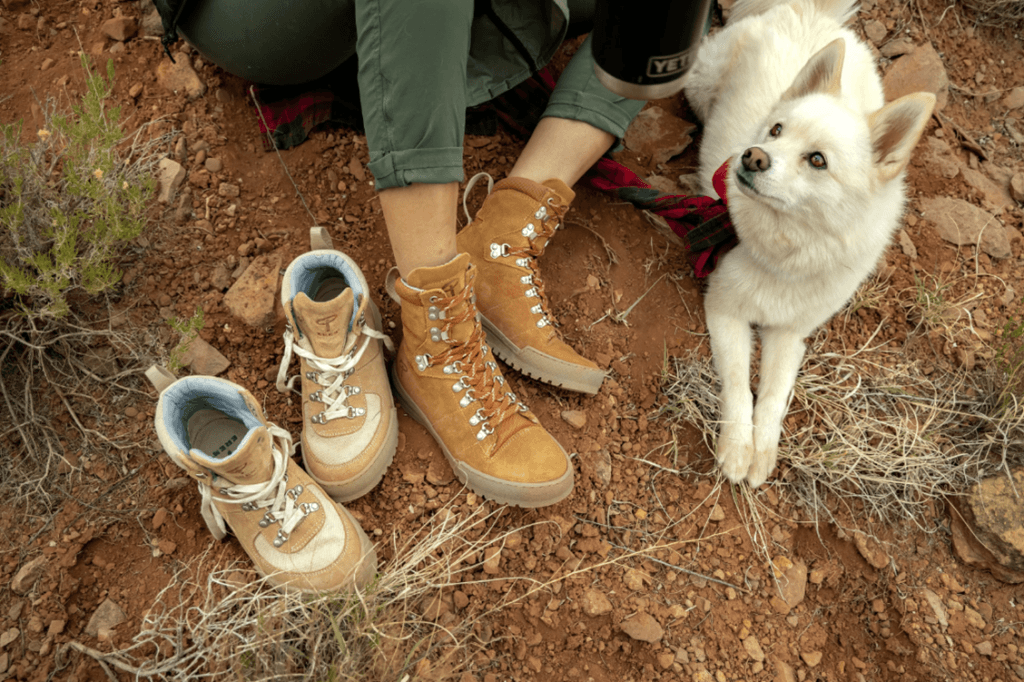 A woman is sitting on the ground with a dog and a pair of hiking boots.
